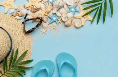 Terlik, güneş gözlüğü ve hasır şapka gibi plaj aksesuarları açık mavi arka planda deniz kabukları ve deniz yıldızlarıyla yatıyordu.