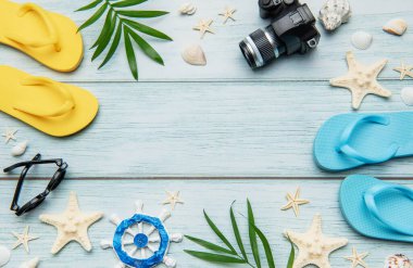 Mavi ahşap arka planda parmak arası terlikler, kamera, güneş gözlüğü, deniz kabuğu, deniz yıldızı ve palmiye yaprakları içeren yaz tatili rahatlama konsepti