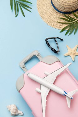 Pembe bavul, oyuncak uçak, güneş gözlüğü, hasır şapka, deniz kabuğu ve mavi arka planda palmiye yaprakları içeren yaz seyahati konsepti.