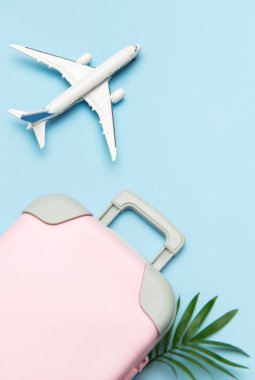 Pembe bavul, oyuncak uçak ve açık mavi arka planda tropikal yaprak yaz tatili planlarını temsil ediyor.