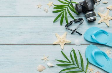 Parmak arası terlikler, kamera, güneş gözlüğü, deniz kabuğu ve palmiye yaprakları mavi ahşap bir arka planda yaz tatili düzenlemesi yapıyor.