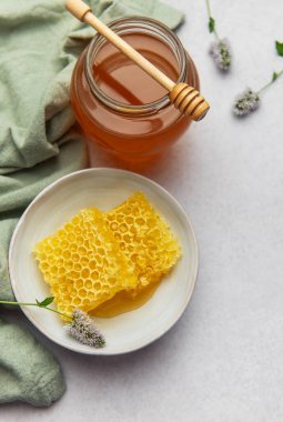 Taze bal peteği ve bal kavanozu ahşap kaplamalı mutfak masasında yeşil bir bez ve narin mor çiçeklerle birlikte oturur.