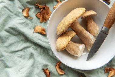 Taze Porcini mantarları ve bıçak kasede, pişmeye hazırlanıyor, yeşil masa örtüsüne saçılmış kurutulmuş mantar dilimleri ile.