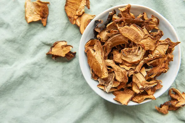 Yeşil bir kumaş üzerinde bir kase kurutulmuş mantarın yukarıdan görünüşü korunmuş mantarların dokusunu ve toprak tonlarını sergiliyor.