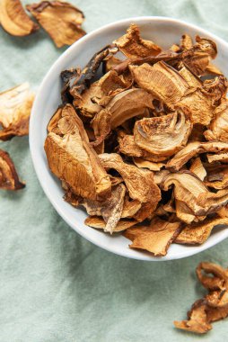 Yeşil bir kumaş üzerinde bir kase kurutulmuş mantarın yukarıdan görünüşü korunmuş mantarların dokusunu ve toprak tonlarını sergiliyor.