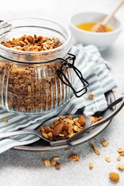 Granola taneleri, fındıklar ve kurutulmuş meyveler gri arka planda ballı bir kavanozda.