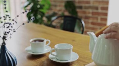 Hand is pouring hot drink from teapot into two tea cups, mugs indoors. Relaxing time at home or in cafe or restaurant. Waiter serving customers. Business with good service. High quality FullHD footage