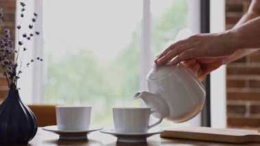 Hand is pouring hot drink from teapot into two tea cups, mugs indoors. Relaxing time at home or in cafe or restaurant. Waiter serving customers. Business with good service. High quality FullHD footage
