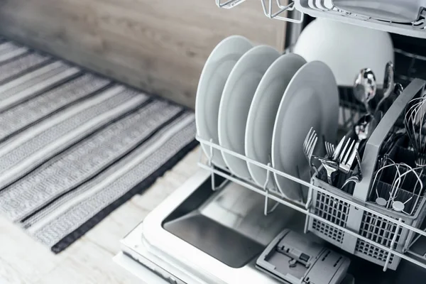 Modern ev mutfağının içinde temiz aletlerle otomatik paslanmaz bulaşık makinesinin kapanışı. Beyaz tabaklar, tabaklar, bardaklar, metal çatallar ve bıçaklar. Ev işleri, ev işleri, ev işleri.