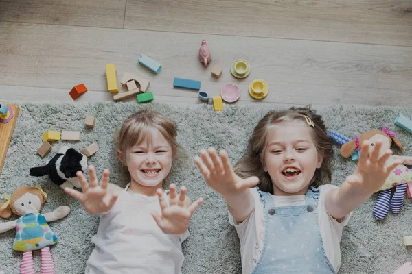 Top view from above on children playing with different toys. Portrait of kids lying on floor at home, kindergarten. Two happy smiling sisters looking at camera with raising hands. Educational games