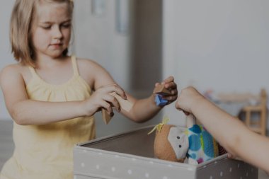 Çocuklar oyun oynadıktan sonra yerden oyuncakları temizliyor, kız kardeş oyuncak bebeklerini tekstil kutusuna toplamaya yardım ediyor. Çocuklar güneşli beyaz ev yatak odasında, anaokulunda ya da çocuk parkında halının üzerinde oturuyorlar.