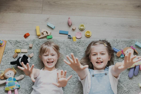 上からのトップビューは 異なるおもちゃで遊んでいる子供たち 幼稚園の床に横たわっている子供たちの肖像画 手を挙げてカメラを見ている2人の幸せな笑顔の姉妹 コンピュータゲーム — ストック写真