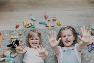Farklı oyuncaklarla oynayan çocukların yukarıdan görünüşü. Evde, anaokulunda yerde yatan çocukların portresi. İki mutlu gülümseyen kız kardeş kameraya el kaldırarak bakıyor. Eğitim Oyunları