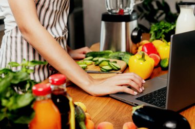Ev mutfağında dizüstü bilgisayar kullanan bir kadın. Kadın el çırpıyor, sebzelerin önünde bilgisayara dokunuyor, masada meyveler var. Seçici odaklanma, kesilmiş. Gıda teslimatı konsepti, mobil diyet uygulaması