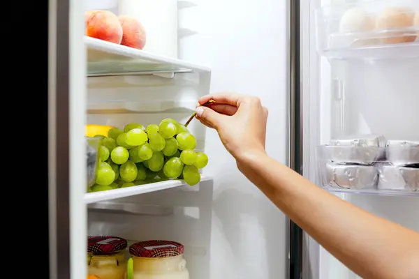 Woman hand taking, grabbing or picks up green bunch of grapes out of open refrigerator shelf or fridge drawer full of fruits, blueberries, bottles with yogurt. Healthy food diet, lifestyle concept