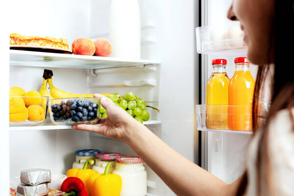 Woman hand taking, grabbing or picks up box of blueberry out of open refrigerator shelf or fridge drawer full of fruits, vegetables, banana, peaches, yogurt. Healthy food diet, lifestyle concept