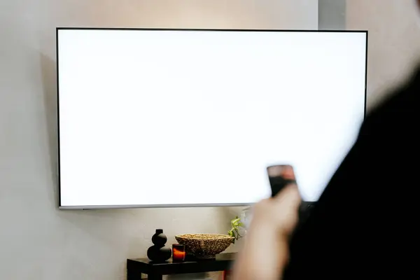 Hand holding remote control pointing to TV. Person watching smart television. Blurred background. White empty mockup screen, copy space. Manage appliances, elements of a smart home control panel
