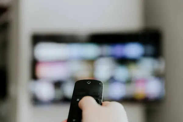 Hand holding remote control pointing to TV. Person watching smart television indoors. Blurred background. Colorful screen. Manage appliances, elements of a smart home control panel, comfort for life