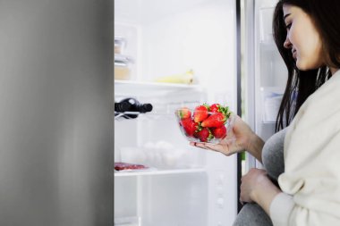 Düşünceli, aç, hamile bir kadın açık buzdolabının önünde duruyor, gecenin bir yarısı mutfaktaki buzdolabından çilek alıyor, kapanıyor. Sağlıklı beslenme, beslenme, hamilelik diyeti