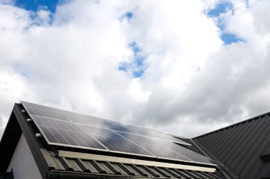 Temiz alternatif enerji. Evdeki metal kaplama çatı katında güneş panelleri olan tek başına fotovoltaik fabrika sistemi olan modern bir ev. Bulutlar, mavi gökyüzü arkaplanı. Profesyonel kurulum, yeşile dön