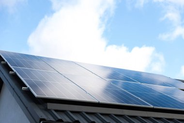 Bulutlu mavi gökyüzü arkaplanındaki güneş paneli hücreleri. Temiz alternatif enerji konsepti. Metal çatıdaki güneş panelleri tek başına fotovoltaik tesis sistemine sahip modern bir ev. Bastır yeşil