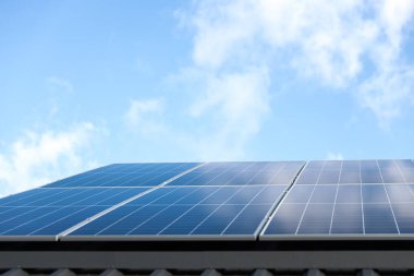 Bulutlu mavi gökyüzü arkaplanındaki güneş paneli hücreleri. Temiz alternatif enerji konsepti. Metal çatıdaki güneş panelleri tek başına fotovoltaik tesis sistemine sahip modern bir ev. Bastır yeşil