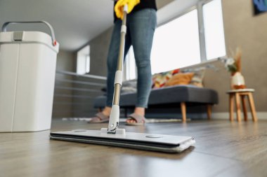 Modern ev ortamında yerleri paspaslayan, parlak ahşap silen bir kadın. Sarı eldiven giyen kız. Ev hanımı profesyonel temizlik malzemesi kullanıyor: paspas, konforlu oturma odasında sıkacağı olan plastik kova.