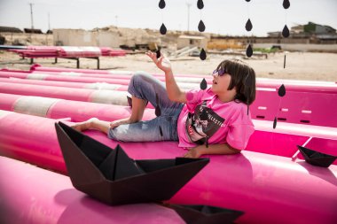 Pembe tişörtlü şirin bir çocuk siyah bulutlu, yağmurlu, mavi gökyüzüne karşı sanayi bölgesinde pembe borularla oynuyor. moda tarzı fotoshoot