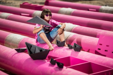 a cute kid in a pink t-shirt with a cat print plays among pink pipes with black clouds, rain, boats and paper planes against the blue sky in an industrial location. fashion style photoshoot