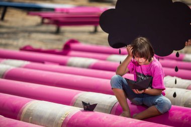 Pembe tişörtlü şirin bir çocuk siyah bulutlu, yağmurlu, mavi gökyüzüne karşı sanayi bölgesinde pembe borularla oynuyor. moda tarzı fotoshoot