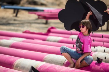 Pembe tişörtlü şirin bir çocuk siyah bulutlu, yağmurlu, mavi gökyüzüne karşı sanayi bölgesinde pembe borularla oynuyor. moda tarzı fotoshoot