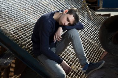 Endüstriyel bir alanda ciddi bir yüzle ve kot pantolonla paslı yapılar ve demir ızgarayla poz veren çekici ve şık bir genç. moda eğilimleri