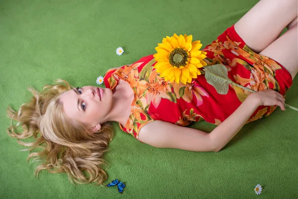 stock image beautiful and sexy long-haired young blonde woman with a charming smile posing barefoot on artificial green grass in a red short dress with print-flowers with a sunflower in her hand 