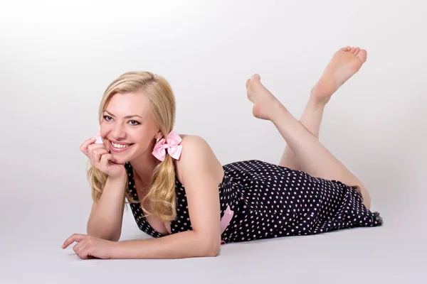 stock image beautiful and sexy fair-haired young woman with a charming smile in a little black dress with white polka dots and a pink belt posing lying down on a light background