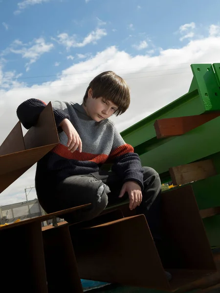 stock image a handsome stylish fair-haired boy posing with a serious face in industry zone among colored iron structures in a knitted sweater and jeans against a bright blue cloudy sky