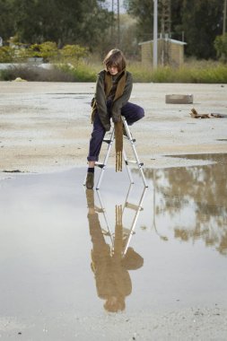 Uzun atkısı ve yağmurlu bir günde ağaçların arka planında bir su birikintisinde merdiven üzerinde poz veren şık kahverengi saçlı beyaz çocuk. moda dergisi konsepti