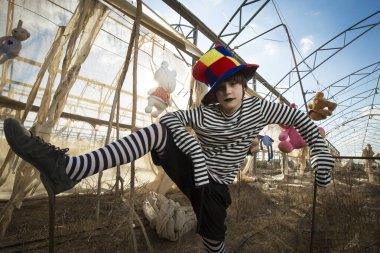 Palyaço kostümü içinde uğursuz bir gülümsemesi olan bir çocuk, mavi bulutlu gökyüzüne karşı, kafeste papağan ve peluş oyuncaklarla terk edilmiş bir serada eğleniyor.