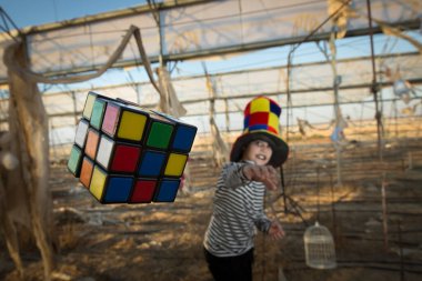 Palyaço kostümü giymiş genç bir adam arka planda dururken bir rubik küp fırlatıyor.