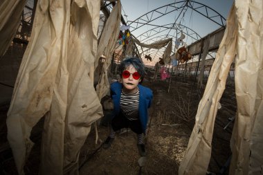 Kırmızı güneş gözlüklü mavi saçlı bir çocuk mavi ceketli ve makyajlı korkunç bir gülümseme terk edilmiş bir serada oyuncakların arasında eğleniyor ve oynaşıyor.