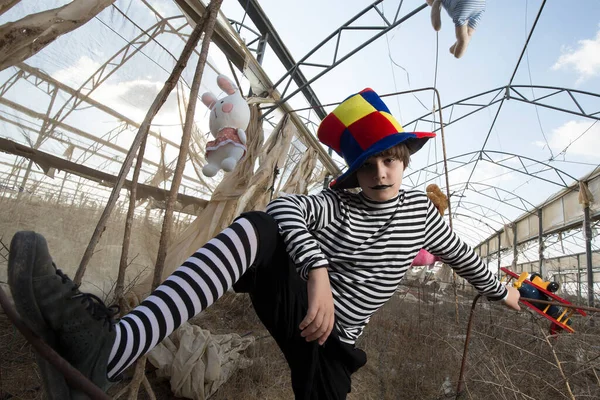 a boy with a sinister smile in a clown costume plays and has fun in an abandoned greenhouse with a parrot in a cage and plush toys, against a blue cloudy sky