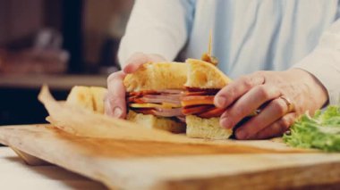 Şef iki dilim sandviçi Fransız ekmeği, peynir, marul ve sosisle açıyor. Kaydırma atışı. Arka planda eski bir fırın tepsisi ve yemek kağıdı var..