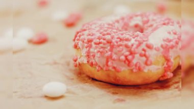 Pembe kremayla süslenmiş pembe çörekler. Çörekler şekerle süslenmiş bir kağıt parçasının üzerinde. Makro ve kaydırma atışı. Şekerler ağır çekimde hareket ediyor. Fırın ve yemek konsepti. Çeşitli renkli çörekler.