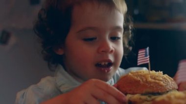 Portrait of American girl with an appetite for biting a hamburger. Young mixed race female eats fast food against a blurred background of a light room. Close up.