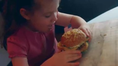 Portrait of American girl with an appetite for biting a hamburger. Young mixed race female eats fast food against a blurred background of a light room. Close up.