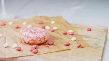 Pembe kremayla süslenmiş pembe çörekler. Çörekler şekerle süslenmiş bir kağıt parçasının üzerinde. Makro ve kaydırma atışı. Şekerler ağır çekimde hareket ediyor. Fırın ve yemek konsepti. Çeşitli renkli çörekler.