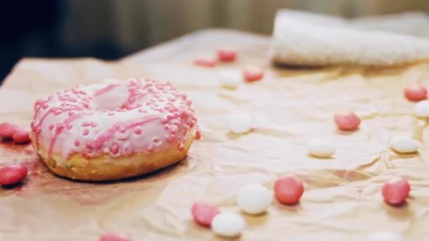 ピンクのドーナツはピンクのアイシングで飾られています ドーナツはキャンディで装飾された紙の上にあります マクロとスライダーの撮影 キャンディーはゆっくりと動きます パン屋と食のコンセプト カラフルなドーナツ — ストック動画