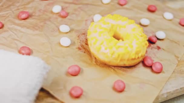 粉红色的甜甜圈装饰着黄色的糖霜 甜甜圈在用糖果装饰的一张纸上 宏观和滑翔机射击 糖果是慢动作的 — 图库视频影像