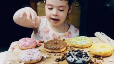 Kız donut yiyor. Çikolata, marşmelov ve retro pişirme tepsisindeki şekerli çörekler. Donutlar doğal çikolatayla süslenmiş bir kağıdın üzerinde. Makro ve kaydırma atışı. Çeşitli renkli çörekler.