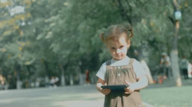Akıllı telefonla oynayan tatlı küçük kız sağlıklı bebek cep telefonuna parmaklarıyla dokunuyor, çizgi film izliyor ve eğitim uygulamalarıyla eğleniyor. Dışarıda parkta bir çocuk