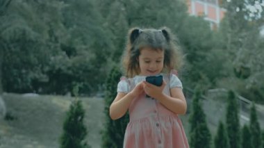 Akıllı telefonla oynayan tatlı bir anaokulu kızı. Sağlıklı 5 yıl. Kız cep telefonuna parmaklarıyla dokunuyor, çizgi film izliyor ve eğitim uygulamalarıyla eğleniyor. Dışarıda parkta bir çocuk